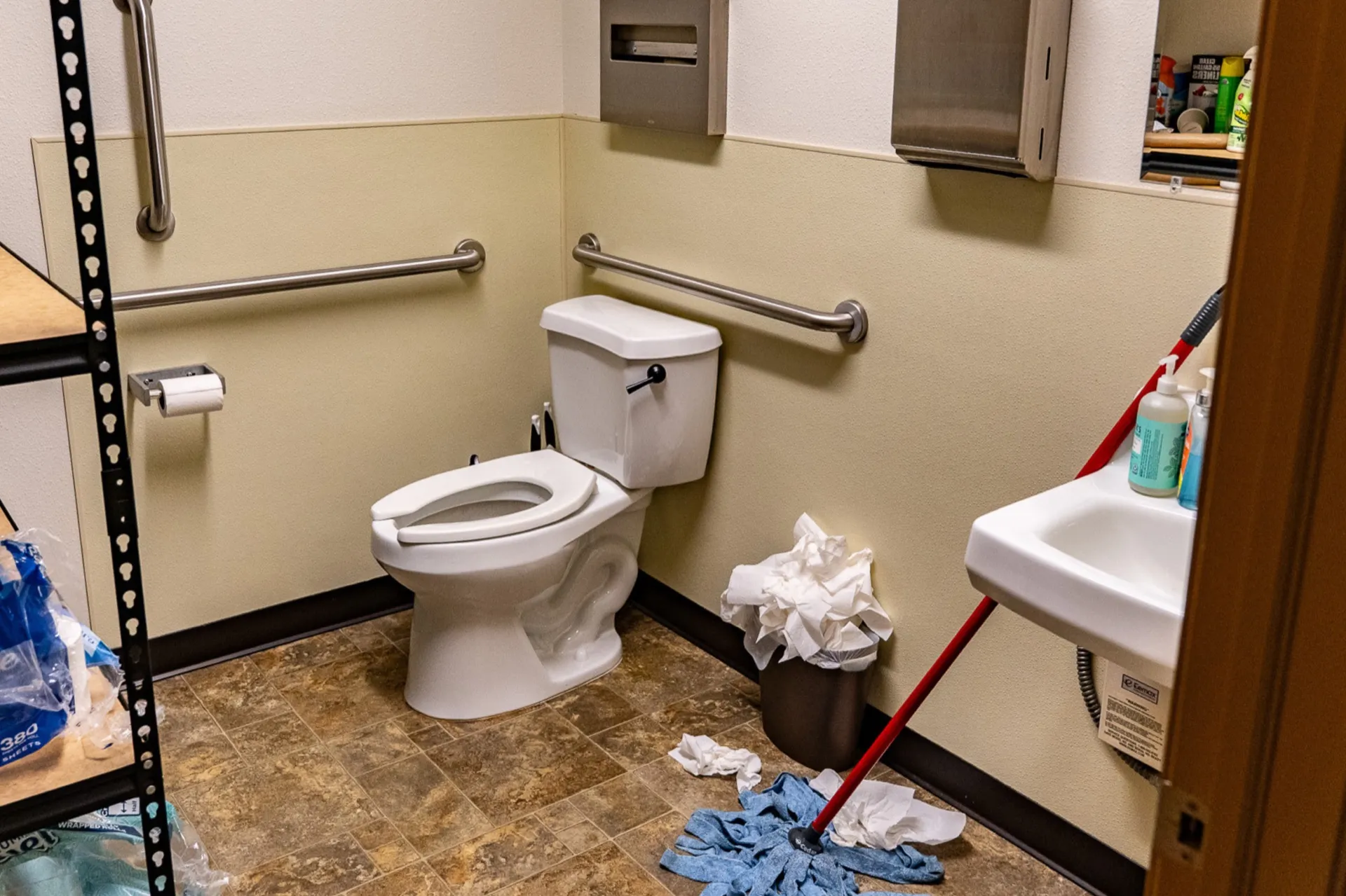 Office restroom before cleaning, with an overflowing trash can and paper towels scattered on the floor