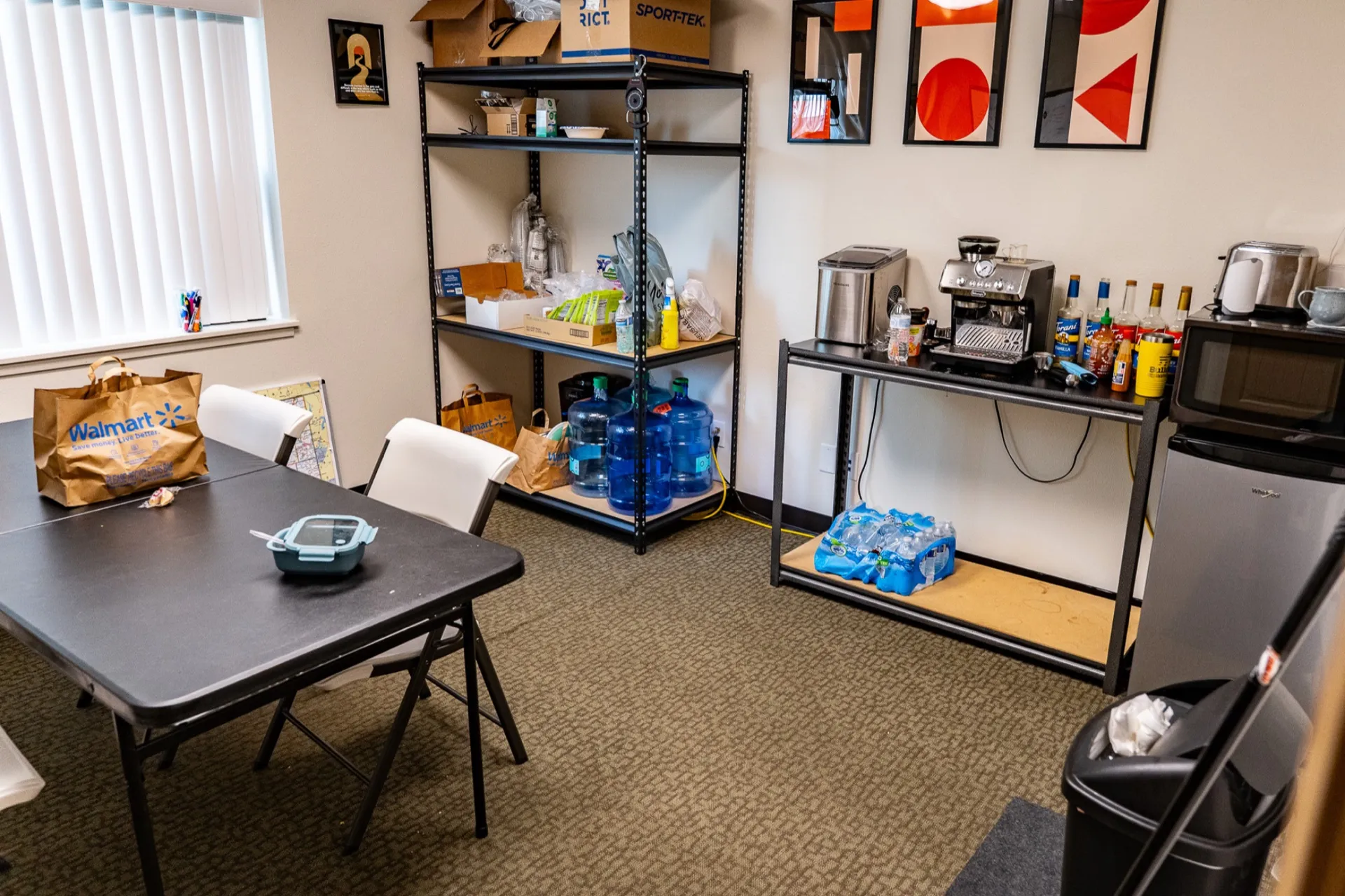 Office break room before cleaning, with shopping bags, clutter, and supplies piled on the table and shelves
